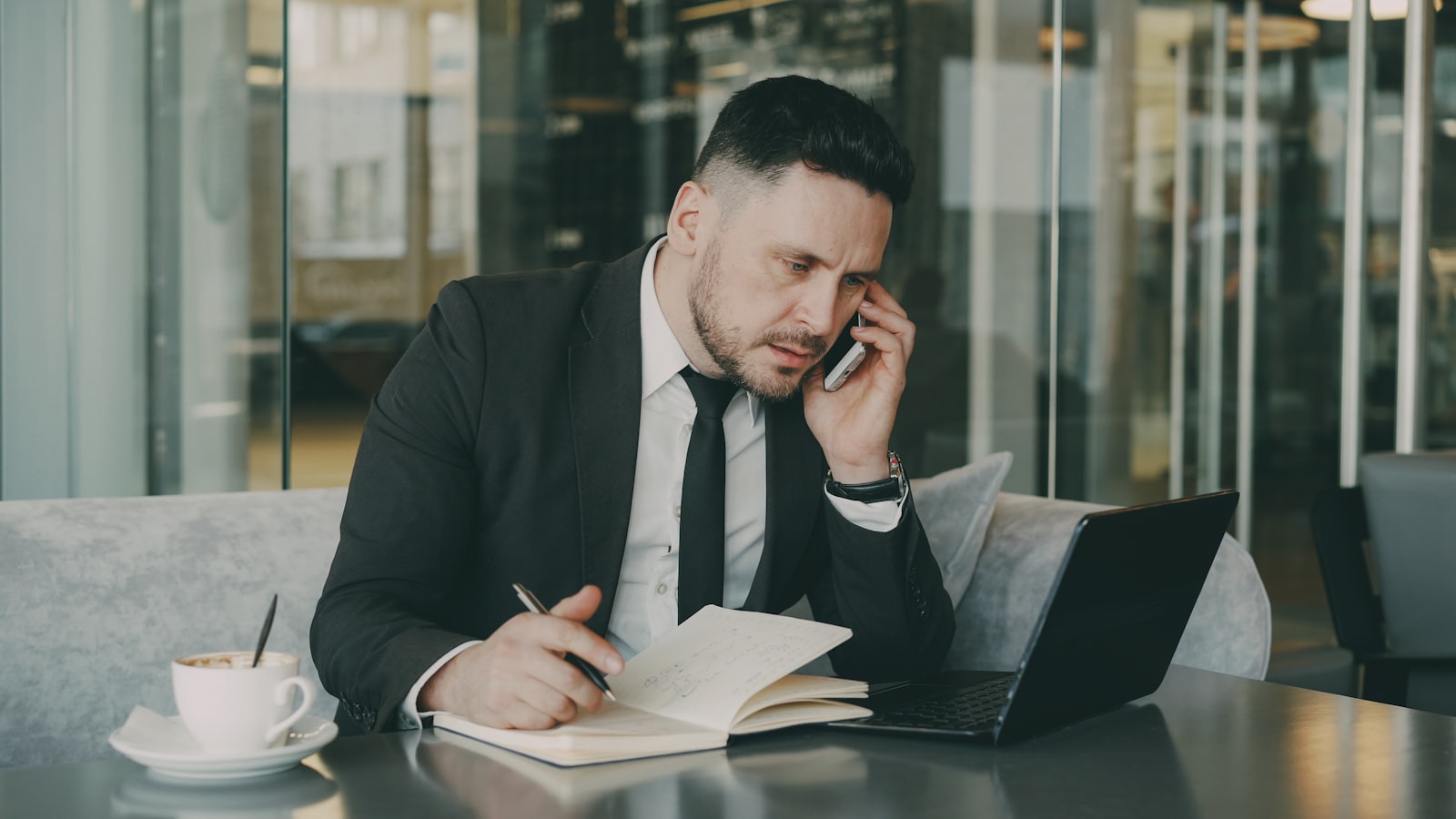 Man in suit talking on phone, writing in notebook.